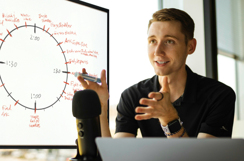 A man sits at a microphone with a dry erase marker in his hand, while his story notes are written on the board behind him.