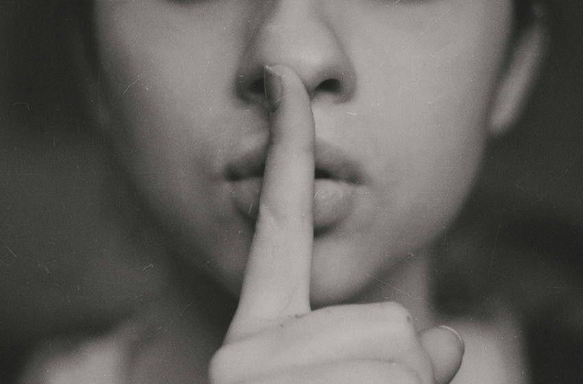 A close up photo of a woman's face as she holds her finger to her lips to signal quiet.