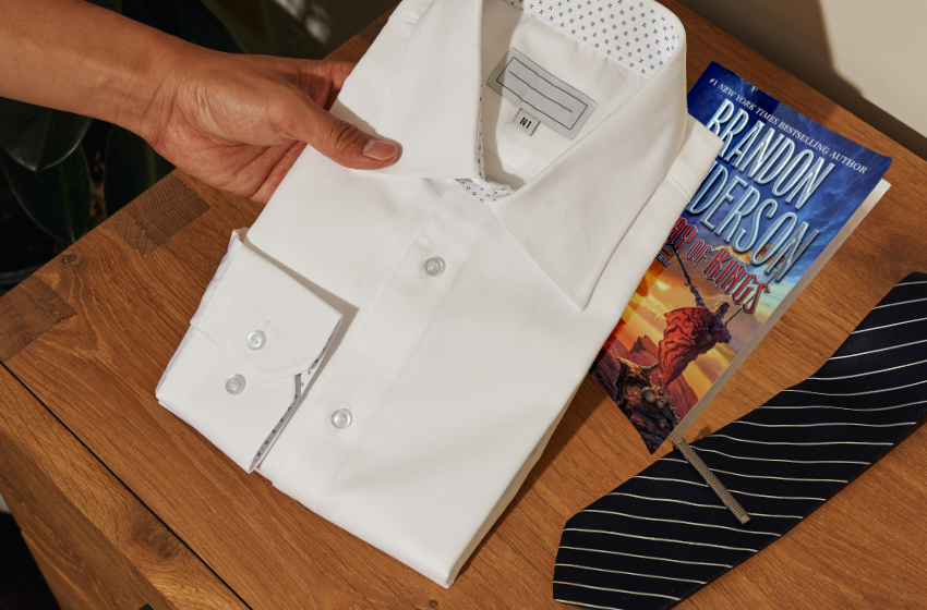 A hand lifts a neatly folded white dress shirt from a table. A tie and a copy of Brandon Sanderson's Way of Kings sit on the table as well.