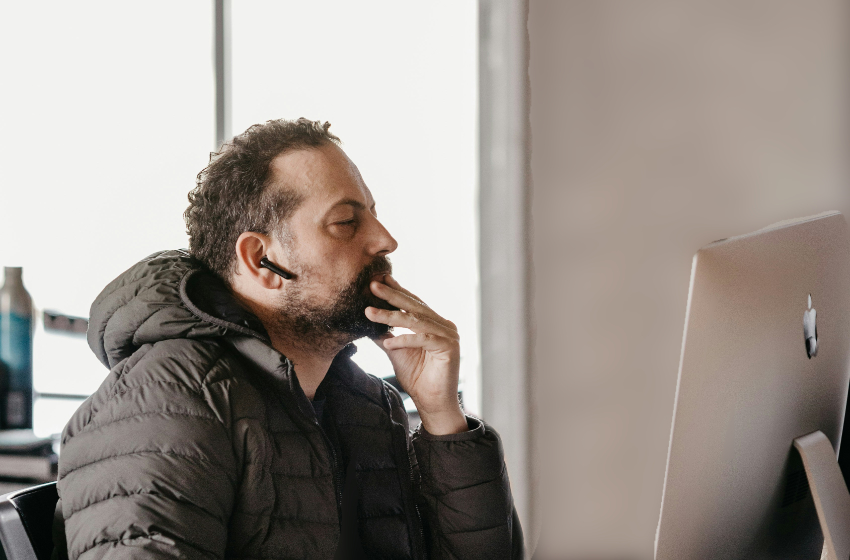 A man with a super cool bluetooth headset stares at a computer screen while rubbing his chin.